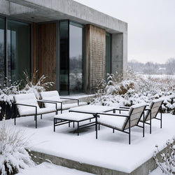 Outdoor lounge set with black steel frames and wooden details covered in snow, placed on a concrete terrace beside a modern Scandinavian home with glass walls and winter landscape.