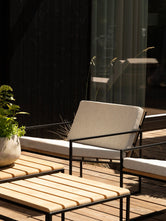 Arvid lounge chair in black steel with light textured cushions, beside teak slat lounge tables in front of the villa