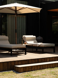 Two Bazo sun loungers in teak and black steel on the wooden deck, with bolster headrests and beige cushions in afternoon light beneath a parasol.