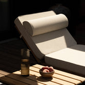 Detail of the Bazo sun lounger with raised backrest and bolster cushion in beige bouclé, beside a teak slat table with a carafe and a bowl of peaches.