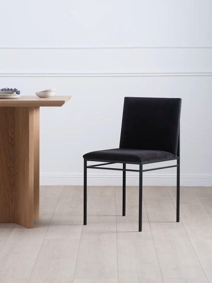 Dining chair Bella in black velvet with a minimalist black frame beside a wooden dining table.