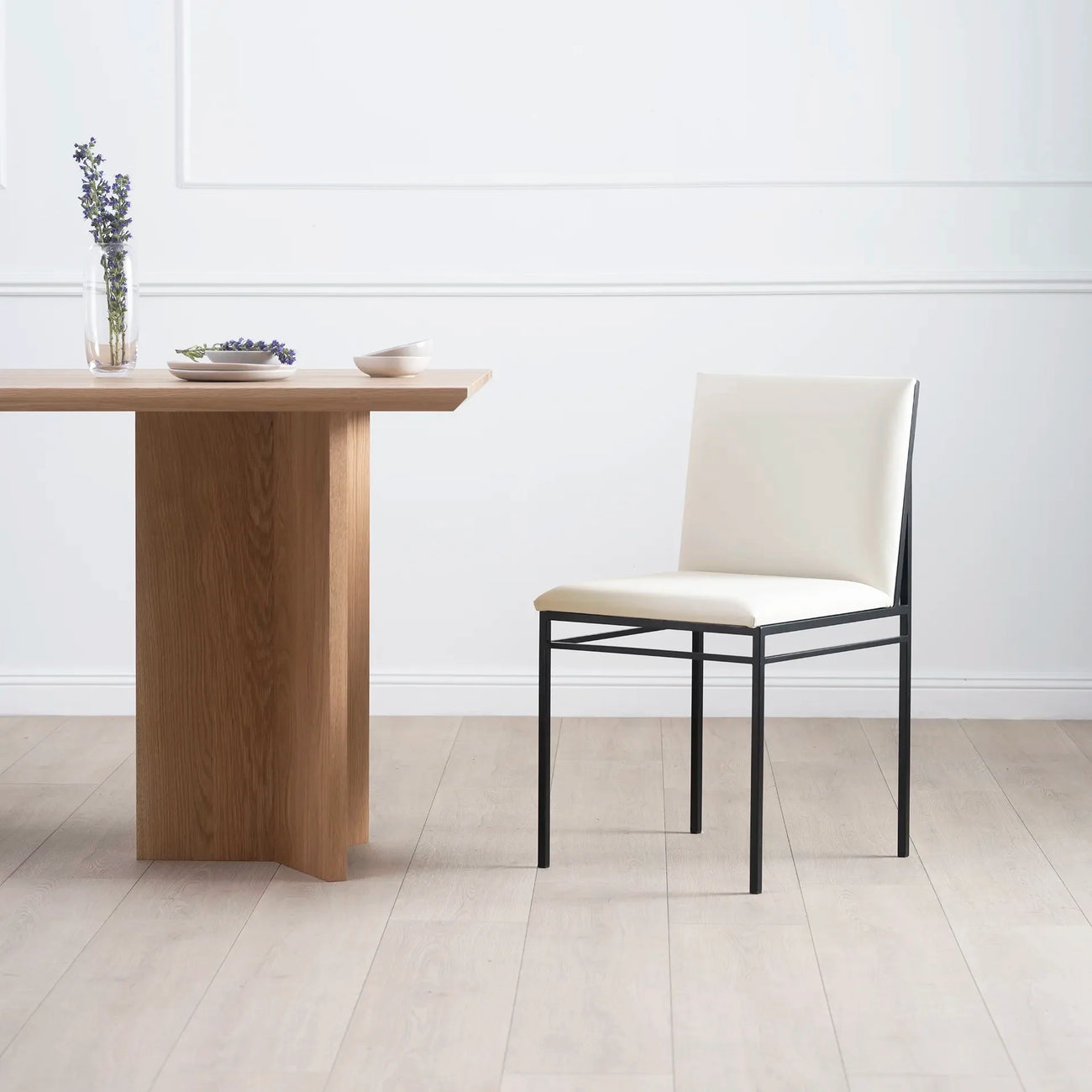 Bella dining chair in ivory leather with a modern black frame, beside a wooden dining table, enhancing minimalist decor.
