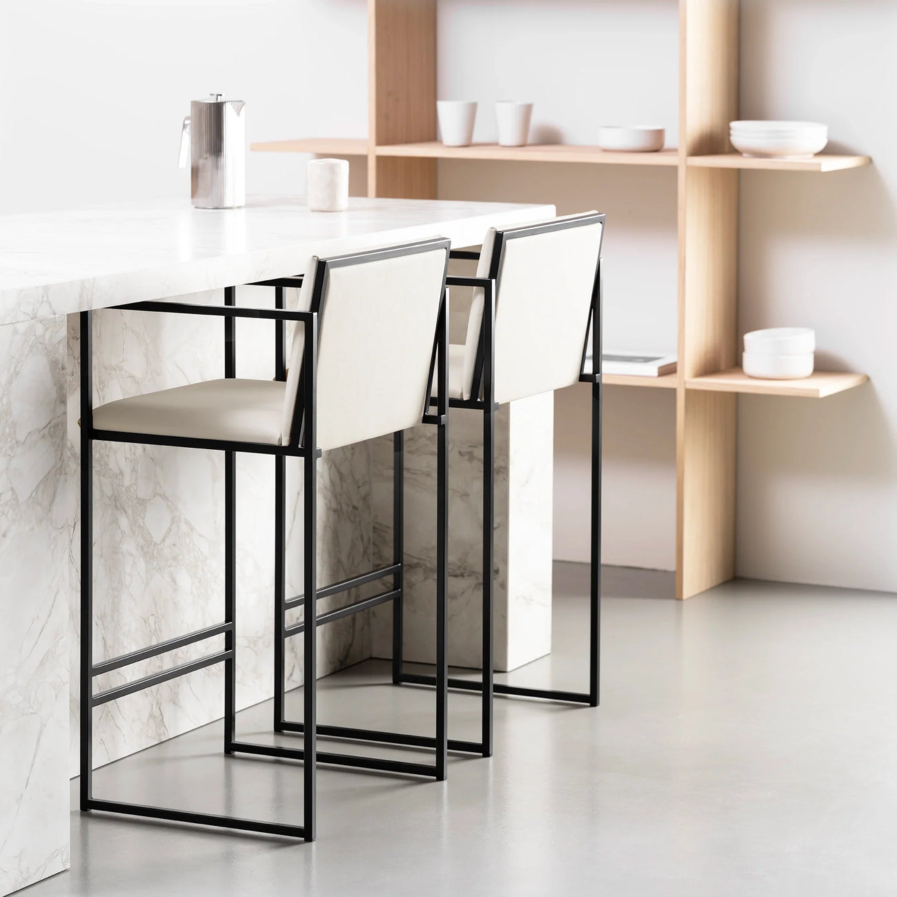 Two Bruno Bar Stools by by Crea with black steel frames and ivory vegan leather upholstery, placed at a white marble kitchen island in a Scandinavian-inspired interior with oak shelving.
