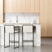 Rear view of two Bruno Bar Stools from by Crea with black steel frames and ivory vegan leather backs, positioned at a white marble kitchen island with natural oak cabinetry.