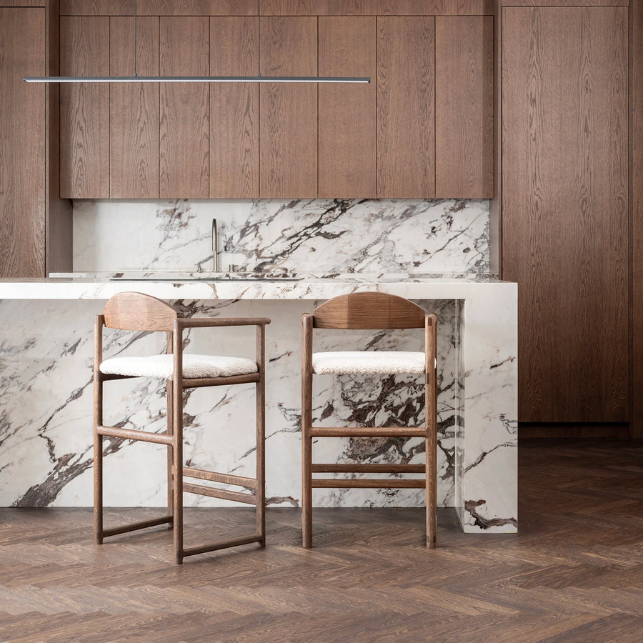 Pair of elegant By Crea Carl bar stools in dark oak with ivory boucle cushions, standing against a luxurious white marble kitchen island and dark wooden cabinetry. Ideal for upscale kitchen designs, Scandinavian furniture, and comfortable seating solutions.