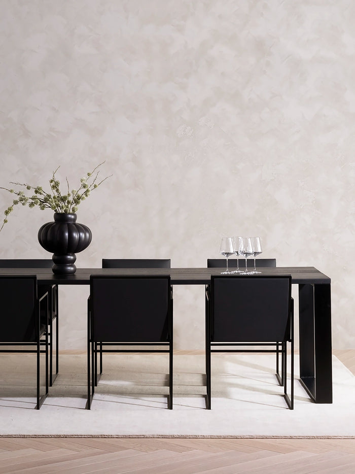 Dining Table Diana in black stained oak with black Sella dining chairs, styled with a black vase against a plaster wall