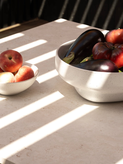 Close-up of the Diana outdoor table’s light Dekton stone sabbia top styled with bowls of peaches, aubergines and tomatoes in sunlight.