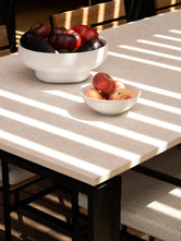 Modern Diana outdoor dining table with a light beige stone-look top and black metal frame, styled with two white fruit bowls as dramatic diagonal shadows create a striking architectural pattern across the tabletop.