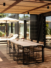 Diana modern outdoor dining table with a light stone top and black frame, paired with Signe teak chairs beneath a slatted pergola.