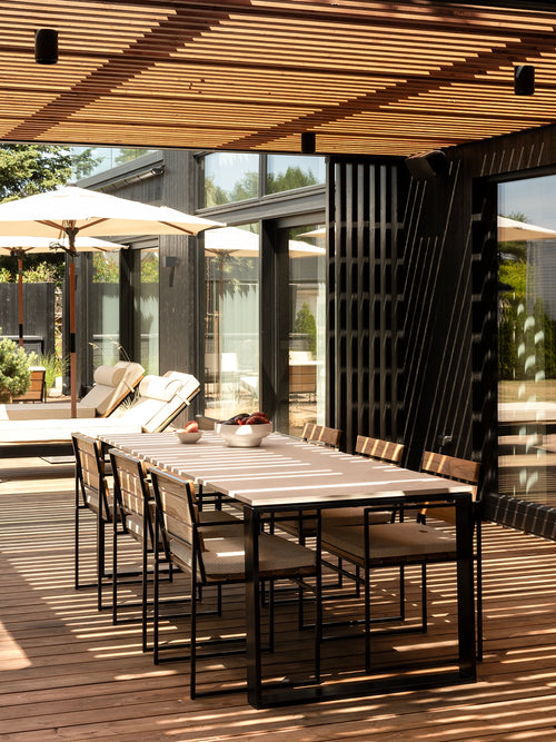 Diana modern outdoor dining table with a light stone top and black frame, paired with Signe teak chairs beneath a slatted pergola.