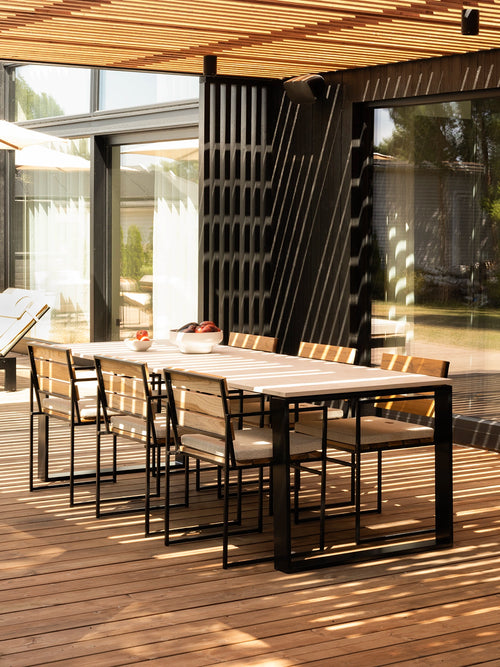 Modern outdoor dining set with a rectangular light wood table and six matching chairs with black metal frames, placed on a wooden deck under a slatted ceiling.