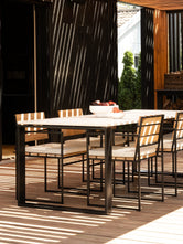 Scandinavian Outdoor dining set with a rectangular table and six matching chairs on a wooden deck, featuring black metal frames and light beige striped cushions, with dappled sunlight and shadows.
