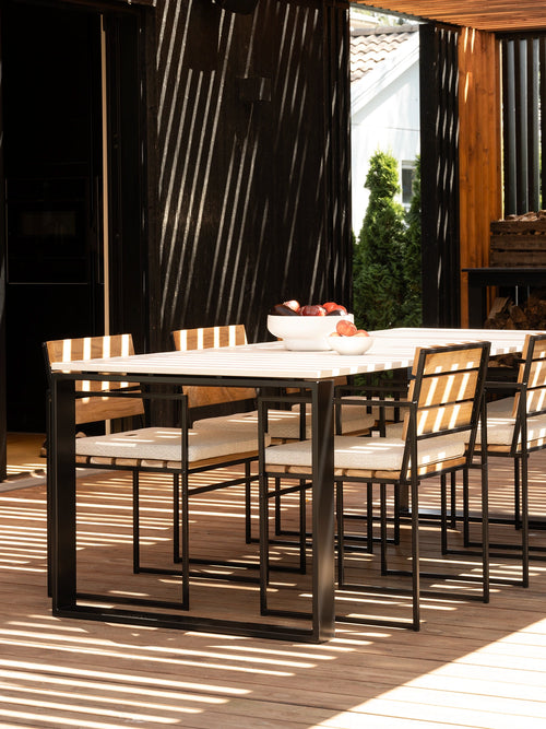 Scandinavian Outdoor dining set with a rectangular table and six matching chairs on a wooden deck, featuring black metal frames and light beige striped cushions, with dappled sunlight and shadows.