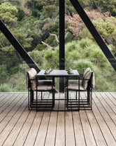 Diana Outdoor Table in sleek black metal paired with four matching armchairs and neutral outdoor cushions, styled on a wooden terrace with panoramic forest views through glass and steel architecture.