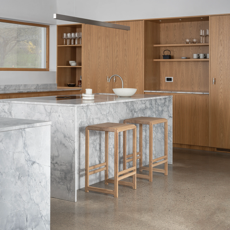 Ebba Bar Stools in solid oak by Crea styled in a Scandinavian kitchen with natural wood cabinetry, grey marble island, and polished concrete floor.