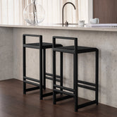 Ebba bar stools in black oak placed at a light stone kitchen island, styled in a warm Scandinavian interior with sheer curtains, dark wood flooring, and minimalist decor.