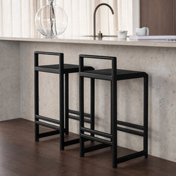Ebba bar stools in black oak placed at a light stone kitchen island, styled in a warm Scandinavian interior with sheer curtains, dark wood flooring, and minimalist decor.
