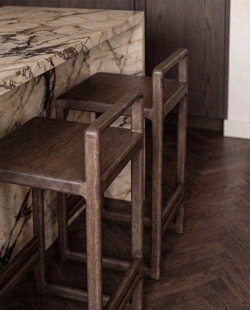 Close-up detail of Ebba bar stools in dark oak tucked under a marble kitchen island, highlighting the solid wood craftsmanship and natural grain texture in a refined Scandinavian interior.