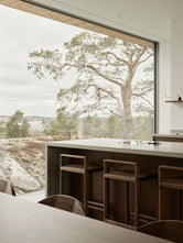 Ebba bar stools in dark stained solid oak at a stone kitchen island, with a full height window framing a pine tree and an archipelago view.