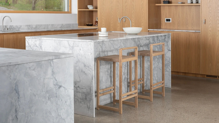 Side view of two Ebba Bar Stools from by Crea in solid natural oak, styled at a marble kitchen island with polished concrete floors and light oak cabinetry in a Scandinavian minimalist kitchen.