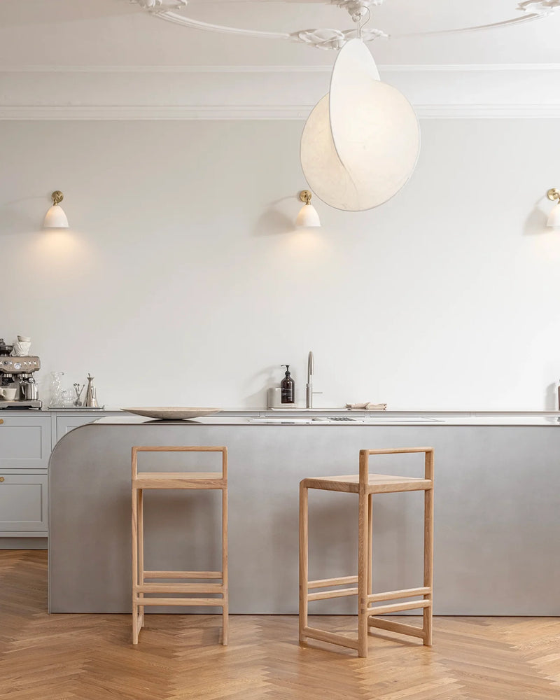 Bright, modern kitchen interior with light grey walls and herringbone parquet flooring, showcasing two Ebba bar stools crafted from light oak wood, with clean lines and footrests, pulled up to a sleek, curved stainless steel or grey-finished kitchen island with a large pendant light overhead.