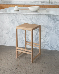 Contemporary kitchen scene featuring a light-toned Eva bar stool made of solid oak, with a simple square seat and sturdy frame, placed against a veined grey marble kitchen island on a concrete floor.