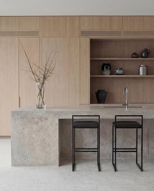Freja bar stools with black steel frames and black upholstered seats placed at a concrete kitchen island, styled in a Scandinavian kitchen with light oak cabinetry and open shelving.