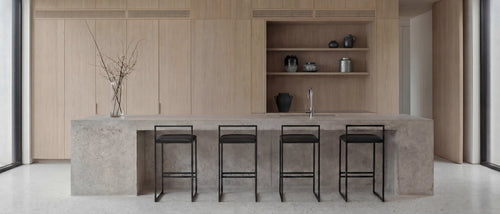 Freja bar stools with black steel frames and upholstered seats placed at a concrete kitchen island, styled in a minimalist Scandinavian kitchen with light oak cabinetry and open shelving.