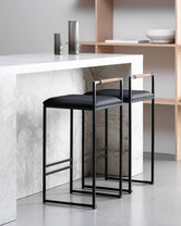 Freja bar stools by by Crea featuring black vegan leather cushions and Coffee Bean wooden backrests, styled at a modern marble kitchen island.