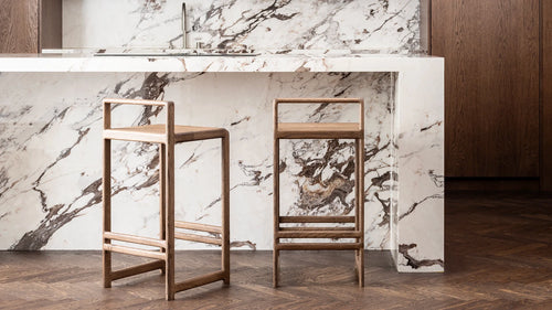 Pair of Ebba Bar Stools in dark solid oak by Crea, styled at a luxurious marble kitchen island with deep wood cabinetry and herringbone floors. Scandinavian design with handcrafted wooden detail.