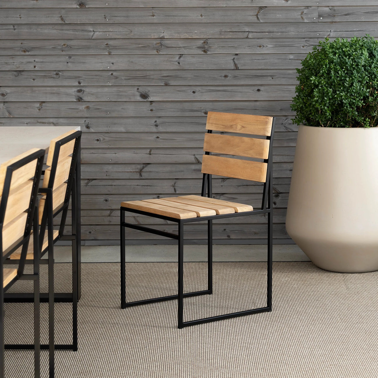 Isa Outdoor Chair by by Crea in teak wood and black steel frame, shown beside a minimalist outdoor dining table on a Scandinavian patio. Custom handmade outdoor furniture for modern living.