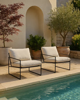 Kenny outdoor lounge chairs with black frame and ivory cushions, styled by a Mediterranean poolside with olive trees and terracotta pots.