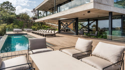 Modern poolside terrace featuring Kenny Outdoor Lounge Chairs with soft beige cushions and a row of Bazo Sunbed Loungers in black frames, arranged on a spacious wooden deck surrounding a luxury swimming pool.