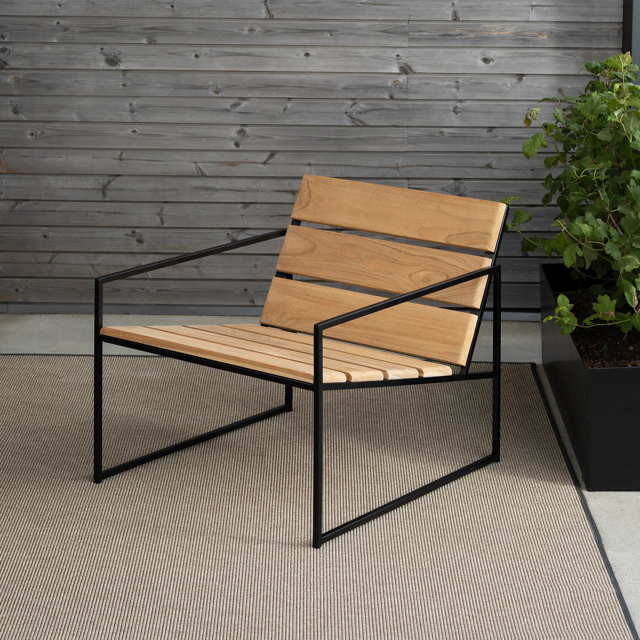 Kenny Outdoor Lounge Chair by by Crea in teak and black frame, shown on a neutral outdoor rug with grey wood cladding and greenery. Handmade Scandinavian furniture for modern patios and terraces.