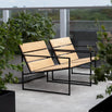 Kenny Outdoor Lounge Chair in teak wood with black steel frame by by Crea, minimalist Scandinavian design. Shown on a modern balcony with lush green plants and concrete flooring, ideal for urban outdoor living, handcrafted in Sweden.