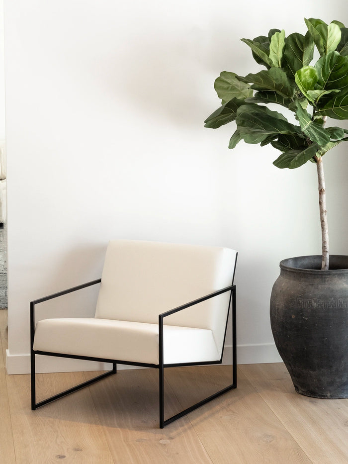 Armchair Kent with black frame and ivory leather cushion beside a potted plant in a modern interior.