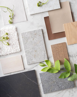 Flatlay of Scandinavian-inspired material samples including oak wood, taupe and brown vegan leather, white bouclé fabric, and Dekton stone surfaces in neutral tones, styled with greenery on a marble background.