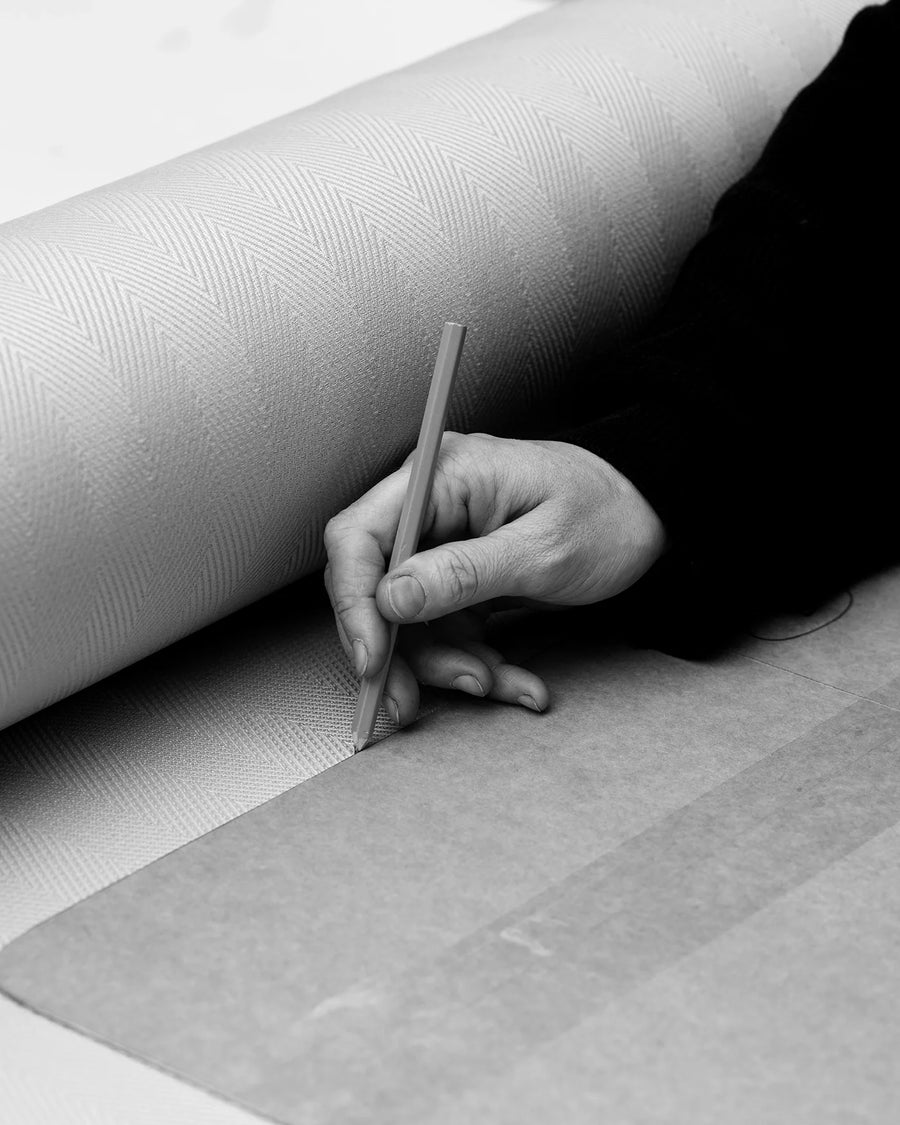 Close-up black-and-white photo of a craftsman's hand precisely tracing fabric patterns on upholstery textile using a pencil and cardboard stencil, representing by Crea’s handmade furniture process.