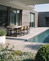Outdoor dining table with black steel frame and wooden chairs placed on a wooden terrace beside a swimming pool, styled in a modern Scandinavian home exterior with natural wood cladding.