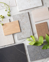 Flatlay of Scandinavian material swatches featuring oak wood in light and medium tones, and Dekton stone surfaces in warm neutrals, soft greys, and deep charcoal, styled with green leaves on a marble backdrop.