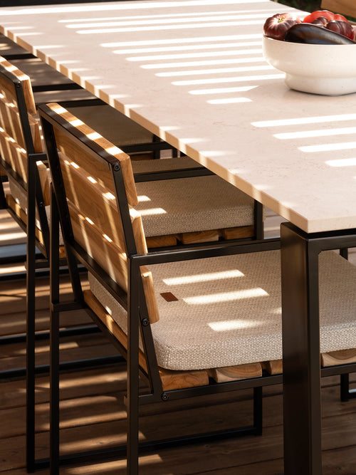 Diana outdoor dining table with a light stone top and dark metal frame, paired with Signe chairs in teak and textured light upholstery, set beneath a slatted pergola where strong sunlight creates graphic striped shadows across the table and terrace floor.