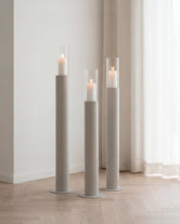 Thomas candleholders in beige with glass cylinders and lit candles, styled on a light wooden floor beside sheer curtains in a calm Scandinavian interior.
