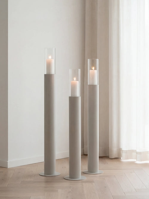 Thomas candleholders in beige with glass cylinders and lit candles, styled on a light wooden floor beside sheer curtains in a calm Scandinavian interior.