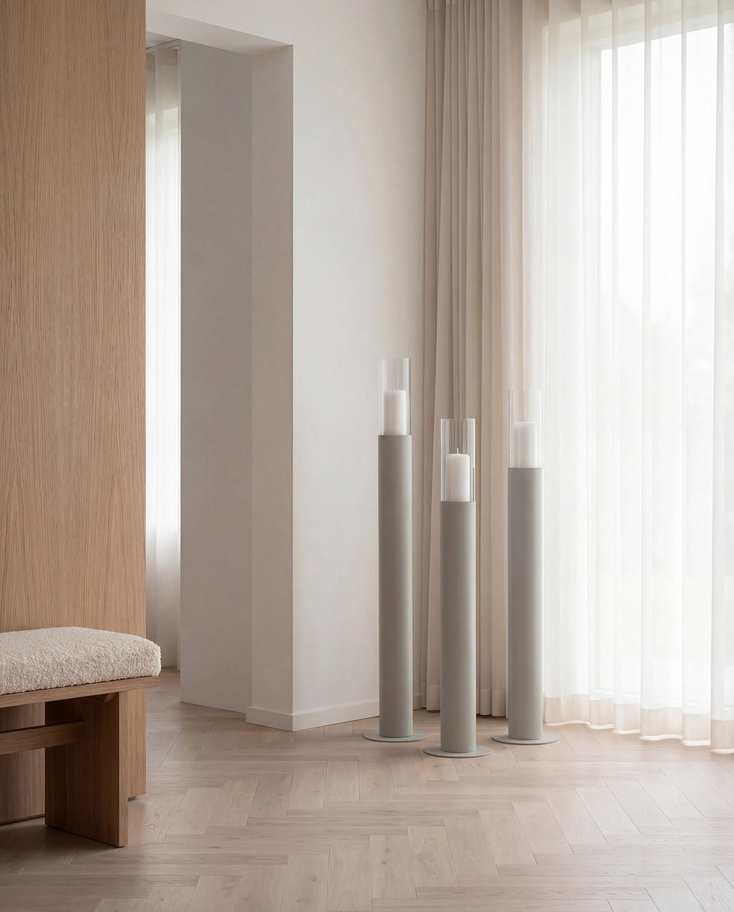 Thomas candleholders in beige with glass cylinders, styled on a light herringbone wooden floor beside sheer curtains in a warm Scandinavian living space with natural wood details.