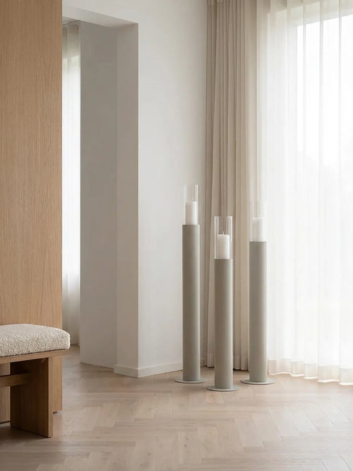Thomas candleholders in beige with glass cylinders, styled on a light herringbone wooden floor beside sheer curtains in a warm Scandinavian living space with natural wood details.