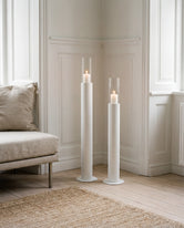 Thomas candleholders in white with glass cylinders and lit candles, styled in a classic Scandinavian interior with wall paneling, light wooden flooring, and a neutral lounge chair.