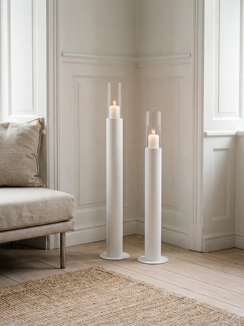 Thomas candleholders in white with glass cylinders and lit candles, styled in a classic Scandinavian interior with wall paneling, light wooden flooring, and a neutral lounge chair.