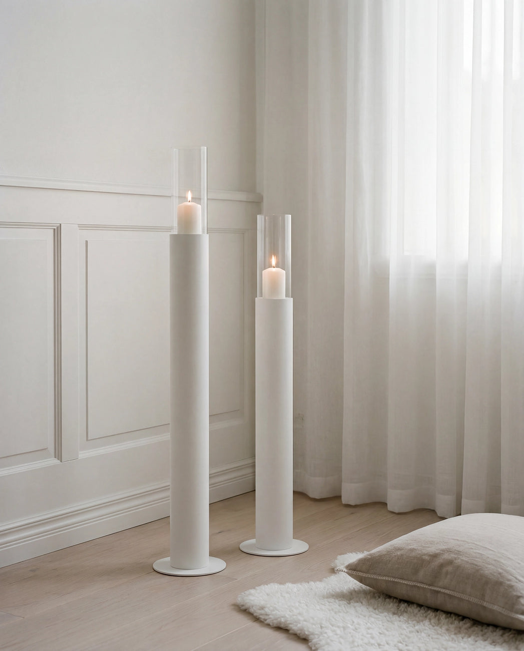 Thomas candleholders in beige with glass cylinders and lit candles, placed on a light wooden floor beside sheer curtains and classic wall paneling in a calm Scandinavian bedroom setting.