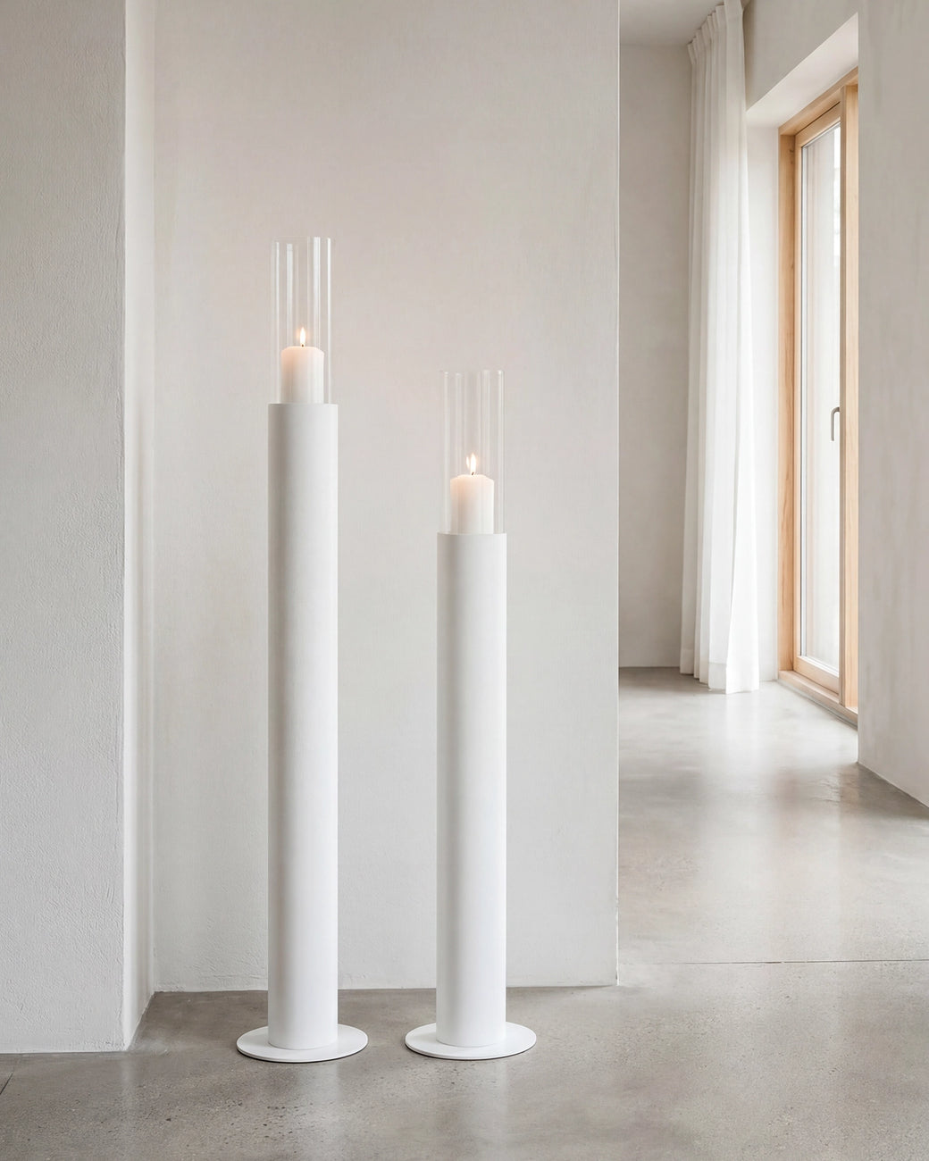 Thomas candleholders in white with glass cylinders and lit candles, placed in a minimalist Scandinavian hallway with concrete flooring, soft white walls, and natural light from wooden-framed windows.