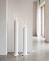 Thomas candleholders in white with glass cylinders and lit candles, placed in a minimalist Scandinavian hallway with concrete flooring, soft white walls, and natural light from wooden-framed windows.